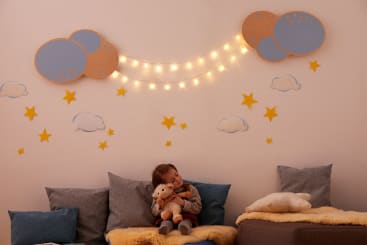 Ein kleines Kind sitzt auf einem Sofa und hält ein Stofftier, umgeben von Kissen und einer gelben Decke. An der Wand dahinter sind dekorative Wolken, gelbe Sterne und Lichterketten zwischen zwei Wolkenformen angebracht.
