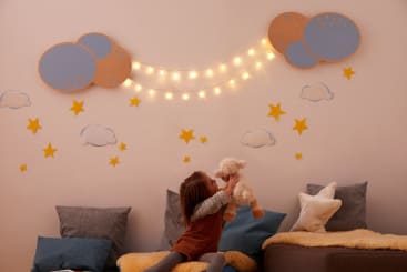 Ein Kind sitzt auf einem Bett und hält ein Stofftier hoch. Die Wand ist mit Wolken, Sternen und Lichterketten in Form von Wolken und einer Girlande geschmückt. Kissen und Decken sind auf dem Bett und dem Boden ausgelegt.