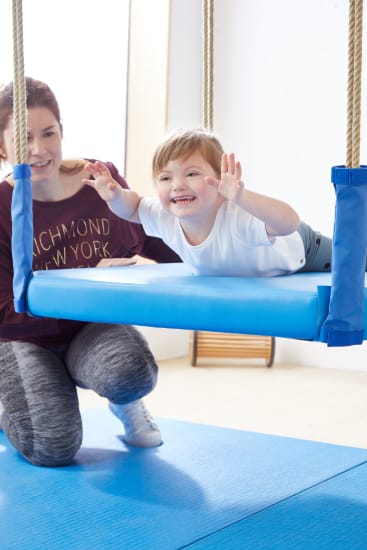 Ein kleines Kind lächelt, während es auf dem Bauch auf einer blauen Schaukel in einer Turnhalle liegt, während ein Erwachsener in der Nähe kniet, um es zu stützen. Beide befinden sich auf blauen Matten in einem hellen Raum.