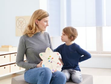 Eine Frau in einem grauen Rollkragenpullover hält eine hölzerne Lehrtafel mit farbigen Wäscheklammern, während sie neben einem Jungen in einem blauen langärmeligen Hemd sitzt. Sie sitzen in einem Raum und schauen sich an.