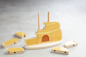 Ein Holzpuzzle in Form eines Schiffes mit mehreren gelben und naturfarbenen Holzteilen, von denen einige auf einem Holzsockel mit zwei vertikalen Stangen montiert sind, während andere Teile auf einer grauen Fläche verstreut liegen.