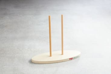 Ein ovaler Holzsockel mit zwei senkrecht stehenden Holzstäben, die auf einer hellgrauen Fläche stehen. Der Sockel hat ein kleines farbiges Logo und einen Text an einer Kante.