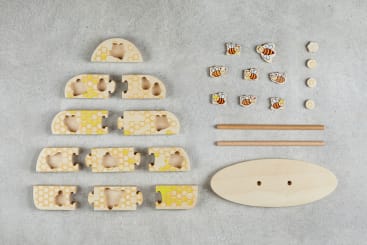 Spielzeugteile für einen Bienenstock aus Holz, die auf einer grauen Fläche ausgelegt sind, einschließlich halbkreisförmiger Bienenstocksegmente mit Wabenmustern, acht Bienenstücken, zwei Holzstäben, einem ovalen Sockel und kleinen runden Verbindungsstücken.