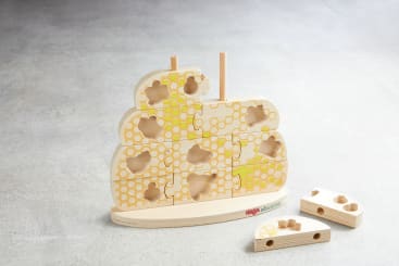 Ein Holzpuzzle in Form eines Bienenstocks mit Bienenwaben und Bienenmotiven. Es fehlen mehrere Teile, zwei Puzzleteile liegen neben dem Puzzle auf dem Boden. Die Marke HABA ist auf dem Sockel sichtbar.