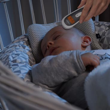 Ein Baby schläft in einem Kinderbett, während jemand ein digitales Thermometer an die Stirn des Babys hält, das einen Messwert anzeigt. Die Bettwäsche für das Kinderbett ist mit einem geometrischen Muster in Grau und Weiß versehen.