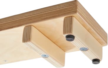 Eine Nahaufnahme der Unterseite eines Holzregals mit zwei Halterungen, die mit Schrauben und schwarzen Gummipads an den Enden befestigt sind. Das Regal und die Halterungen sind aus hellem Sperrholz gefertigt.