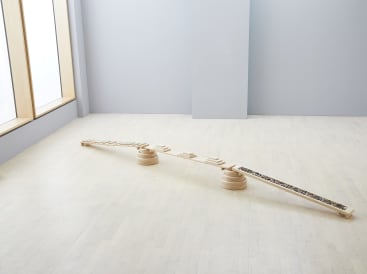 Eine minimalistische Installation in einer hellen Galerie zeigt einen langen Holzbalken, der auf zwei runden Holzstützen balanciert, mit kleinen rechteckigen Blöcken und einem Abschnitt, der mit dunklen Steinen oder Kies gefüllt ist.