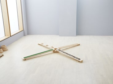 Ein minimalistischer Raum mit hellem Holzfußboden enthält einen X-förmigen Balancierbalken aus Holz. Jeder Balken weist unterschiedliche Texturen oder Oberflächen auf, darunter grün, glatt und ein Abschnitt mit Steinen. Durch große Fenster auf der linken Seite fällt natürliches Licht ein.