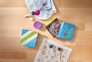 Ein Holztisch mit Bastelmaterial, darunter eine Farbpalette, Schwammspachtel, Glitter, eine Schablone mit Sternformen, bunte Streusel, verzierte Schaumstoffquadrate und Tuben mit metallischer Glitterfarbe.