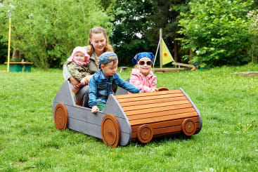Eine Frau und drei kleine Kinder sitzen zusammen in einem hölzernen Spielgerät, das wie ein Auto geformt ist, auf einer Wiese mit Bäumen und Spielgeräten im Hintergrund.