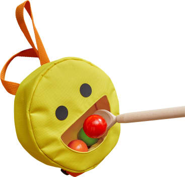 Eine gelbe, runde Plüschtasche mit schwarzen Augen und einem offenen Mund, der an einen Smiley erinnert, mit einem Holzstab, der rote und grüne Kugeln in die Mundöffnung steckt. Ein orangefarbenes Band ist an der Tasche befestigt.