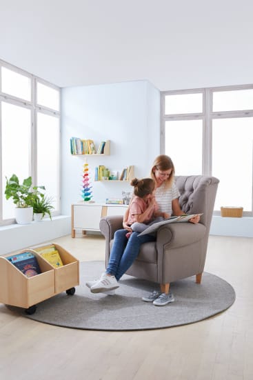 Ein Erwachsener und ein Kind sitzen zusammen auf einem grauen Sessel und lesen ein Buch in einem hellen Raum mit großen Fenstern, Holzböden, einem runden Teppich, Bücherregalen, einer Topfpflanze und einem rollenden Bücherwagen.