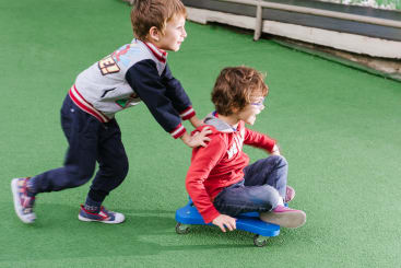 Zwei kleine Kinder spielen draußen auf einem grünen Bodenbelag. Ein Kind sitzt auf einem kleinen blauen Brett mit Rädern, während das andere es von hinten schiebt. Beide tragen legere Kleidung und scheinen Spaß an der Aktivität zu haben.