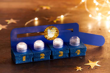 Ein blauer, glitzernder Adventskranz mit vier batteriebetriebenen Teelichtern, die jeweils auf einem von 1 bis 4 nummerierten Feld stehen, wird auf einer Holzfläche mit sternförmigen Dekorationen und Lichterketten im Hintergrund aufgestellt.