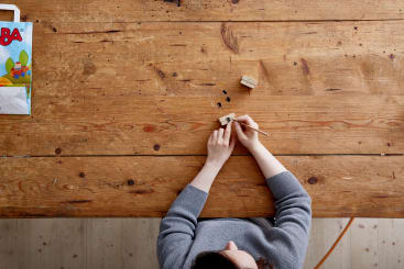 Eine Person, von oben gesehen, hält einen Bleistift und zeichnet auf einem kleinen Holzblock an einem Holztisch. Die Tischoberfläche weist Kratzer und Flecken auf. Eine teilweise sichtbare bunte Schachtel befindet sich in der oberen linken Ecke.