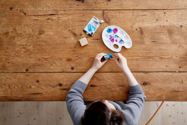 Eine Person sitzt an einem Holztisch und hält eine Farbtube in der Hand. Auf dem Tisch befinden sich eine Farbpalette mit blauer und magentafarbener Farbe, eine weitere Farbtube und ein kleiner Holzblock. Die Szene wird von oben betrachtet.