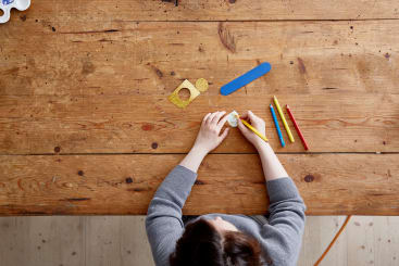 Eine dunkelhaarige Person sitzt an einem Holztisch und zeichnet mit einem gelben Bleistift auf einen kleinen runden Gegenstand. Auf dem Tisch liegen vier Buntstifte, ein blaues Lineal und geometrische Schablonen. Die Szene wird von oben betrachtet.