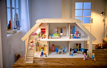 Ein hölzernes Puppenhaus mit zwei Etagen steht auf einem Tisch. Es ist mit Miniaturbetten, -tischen und -stühlen möbliert, und mehrere kleine Puppen befinden sich in verschiedenen Zimmern, in denen verschiedene Aktivitäten dargestellt werden.
