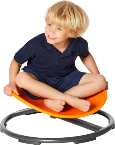 Ein kleines Kind mit blondem Haar, marineblauem Hemd und Shorts, sitzt im Schneidersitz auf einem orangefarbenen, runden Wackelstuhl mit grauem Sockel vor weißem Hintergrund.