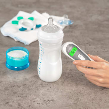 Eine Hand hält ein digitales Thermometer neben einer mit Milch gefüllten Babyflasche. In der Nähe stehen ein Behälter mit Puder, gefaltete Tücher, ein Schnuller und ein Beißring auf einer grauen Arbeitsplatte.