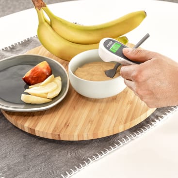 Eine Hand hält ein digitales Lebensmittelthermometer über eine Schüssel mit Babynahrung auf einem Holzbrett. Auf dem Brett, das auf einem Tisch mit einem grauen Tischset steht, befinden sich auch geschnittene Apfelstücke und zwei Bananen.