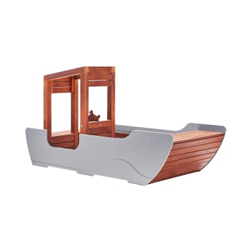 Ein Kinderspielgerät aus Holz, das einem Boot nachempfunden ist, mit einem grauen Rumpf, einem braunen Lattendeck und einer kleinen erhöhten Kabine mit einem offenen Fenster und einem dekorativen Holzsteuerrad.