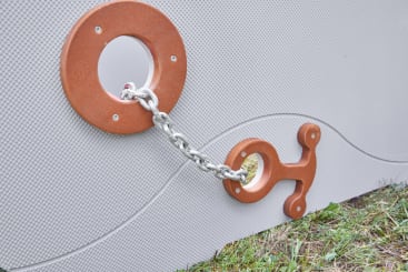 Eine Metallkette verbindet zwei braune runde Leuchten, die an einer grauen Wand mit Wellenmuster angebracht sind; die rechte Leuchte hat eine dekorative, ankerartige Form. Unten rechts auf dem Bild ist Gras zu sehen.