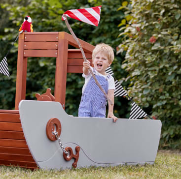Ein kleines Kind steht in einem hölzernen Spielgerät in Form eines Bootes und hält einen Stock mit einer rot-weiß gestreiften Flagge. Auf dem Dach befindet sich ein Spielzeugpapagei und auf dem Boot sind schwarz-weiße Wimpel angebracht. Im Hintergrund sind Bäume zu sehen.