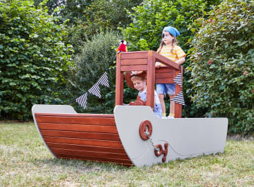 Zwei Kinder spielen auf einem hölzernen Spielgerät in Form eines Schiffes in einem grasbewachsenen Hof; eines steht am Steuerrad des Schiffes und trägt ein blaues Halstuch, während das andere von unten auf das Deck schaut. Sie sind von Bäumen und Büschen umgeben.