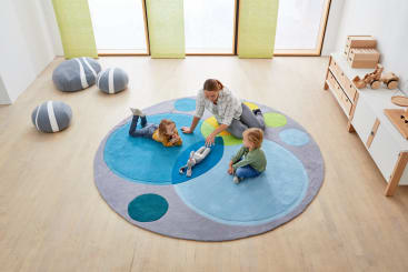 Ein Erwachsener und zwei kleine Kinder sitzen und liegen auf einem kreisförmigen, bunten Teppich mit blauen und grauen Kreisen in einem hellen Raum mit hellem Holzboden, Spielzeugablage und weichen grauen Hockern. Auf dem Teppich liegt ein ausgestopftes Kaninchenspielzeug.