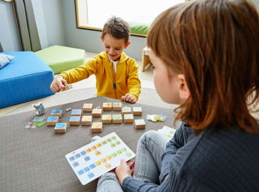 Zwei Kinder sitzen an einem Tisch und spielen ein Zuordnungsspiel mit quadratischen Spielsteinen und bunten Karten. Ein Kind in einer gelben Jacke lächelt und greift nach einem Spielstein, während das andere Kind eine Karte hält und beobachtet.