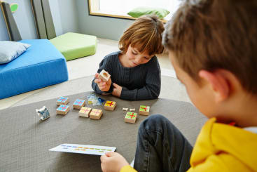 Zwei Kinder sitzen auf dem Boden und spielen ein Lernspiel mit Holzklötzen und einer Karte. Ein Kind hält einen Klotz, während das andere auf die Karte schaut. Im Hintergrund sind helle Kissen zu sehen.