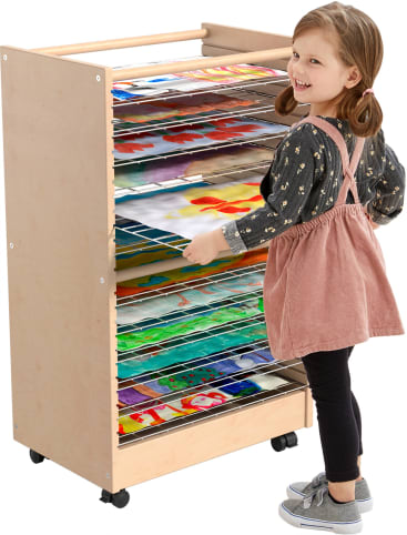 Ein kleines Kind steht neben einem hölzernen Wäscheständer, der mit bunten Kunstwerken gefüllt ist, die auf mehreren Drahtregalen ausgestellt sind. Das Kind lächelt und hält einen der Trockenständer in der Hand.