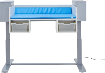 Ein weiß-grauer Wickeltisch mit einer blauen gepolsterten Matte auf der Oberseite, zwei offenen Ablagefächern darunter und einem Stromkabel, das auf der rechten Seite des Rahmens sichtbar ist.