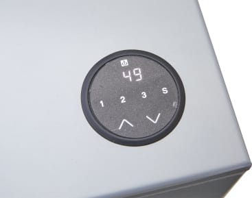 Eine Nahaufnahme eines runden, schwarzen elektronischen Bedienfelds auf einer grauen Fläche zeigt die Zahl 49 auf einem kleinen Bildschirm an. Unterhalb des Bildschirms befinden sich Tasten mit den Bezeichnungen 1, 2, 3, S sowie Auf- und Abwärtspfeile.
