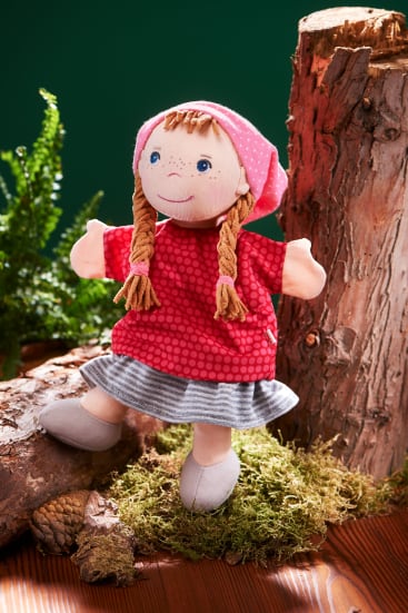 Eine Stoffpuppe mit geflochtenem blondem Haar, rosa Kopftuch, rotem Kleid und grau gestreiftem Rock steht auf moosbewachsenem Boden zwischen Baumstämmen, mit Farnen im Hintergrund.