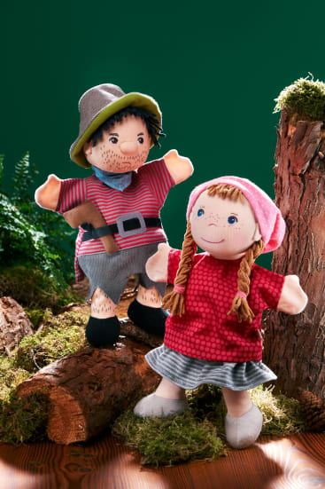 Zwei Handpuppen stehen auf einer Holzfläche mit Moos und Baumstümpfen. Eine ist als Mann mit gestreiftem Hemd, Hut und Gürtel gekleidet, die andere als Mädchen mit Zöpfen, rotem Kleid und rosa Kopftuch. Grüner Hintergrund.