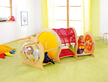 Zwei kleine Kinder spielen auf einem Klettergerüst aus Holz und Plastik in einem hellen Raum mit grünem Boden, gelben Vorhängen und großen Fenstern. Ein Kind krabbelt in einem Tunnel, während das andere am gegenüberliegenden Ende sitzt.