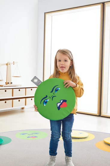 Ein kleines Kind steht in einem Haus und hält ein großes, rundes, grünes Kissen in der Hand, auf dem ein unglückliches Gesicht aufgedruckt ist. Weitere ähnliche Kissen mit unterschiedlichen Ausdrücken liegen in der Nähe auf dem Boden.