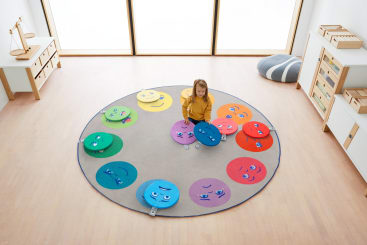 Ein junges Mädchen sitzt auf einem runden Teppich mit bunten, sich überlappenden Kreisen mit Gesichtern, jedes in einer anderen Farbe, in einem geräumigen Klassenzimmer mit hellem Holzboden und Regalen.