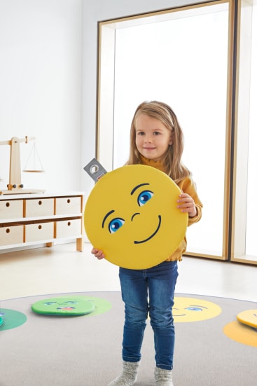 Ein kleines Kind steht in einem Raum und hält ein großes, rundes gelbes Kissen mit einem lächelnden Gesicht in der Hand. Der Raum hat natürliches Licht, Holzmöbel und weitere Kissen auf dem Boden mit ähnlichen Gesichtsmotiven.
