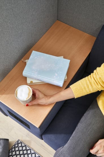 Eine Person in einem gelben Pullover sitzt auf einem grauen Stuhl und hält eine Tasse Cappuccino über einen hellen Holztisch. Zwei geschlossene Bücher, eines mit hellblauem Einband, liegen auf dem Tisch. Ein gemusterter Teppich ist teilweise sichtbar.