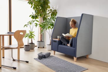 Eine Person sitzt im Schneidersitz auf einem gepolsterten Stuhl mit hoher Rückenlehne und liest ein Buch. Neben ihr steht ein Kaffeebecher. In einem hellen Raum mit Holzfußboden stehen mehrere Topfpflanzen, ein Tisch und ein Teppich.