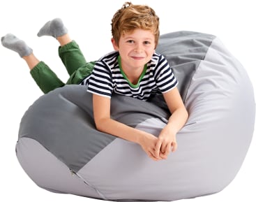 Ein Junge mit hellbraunem Haar, der ein gestreiftes Hemd, eine grüne Hose und graue Socken trägt, liegt auf dem Bauch auf einem großen grauen Sitzsack und lächelt in die Kamera.