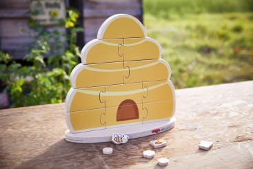 Ein bienenstockförmiges Holzpuzzle mit mehreren fehlenden Teilen liegt auf einem Holztisch im Freien. Einige Puzzleteile, darunter eines mit einer Bienenillustration, sind vor dem Puzzle platziert. Im Hintergrund sind Gras und eine Holzkonstruktion zu sehen.