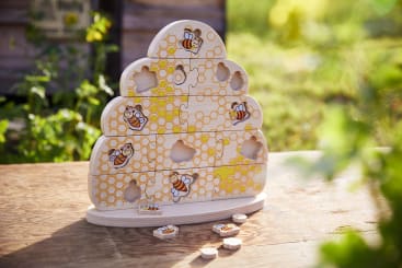 Ein Holzpuzzle in Form eines Bienenstocks mit Bienenwaben und Bienenmotiven liegt auf einer Holzfläche im Freien. Mehrere Teile werden entfernt, wodurch Lücken im Puzzle sichtbar werden. Im Hintergrund erscheinen grünes Laub und Sonnenlicht.