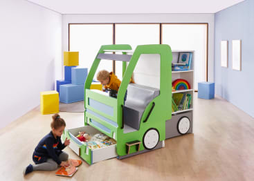 Zwei Kinder spielen in einem hellen Raum mit einem grün-weißen Spielgerät in Form eines Lastwagens, der über Regale und Ablagen verfügt. Ein Kind sitzt auf dem Boden und liest, das andere lehnt sich aus dem Inneren des Lkw-Geräts heraus.