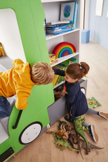 Zwei Kinder spielen neben einem grünen Bücherregal, das wie ein Fahrzeug geformt ist, und greifen nach Dinosaurierspielzeug und Büchern in den Holzregalen, während weiteres Dinosaurierspielzeug auf dem hellen Holzboden verstreut ist.