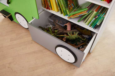 Eine offene Schublade in Form eines Fahrzeugs ist mit Dinosaurierspielzeug aus Plastik gefüllt und ist Teil eines Bücherregals mit Kinderbüchern auf einem Holzboden.