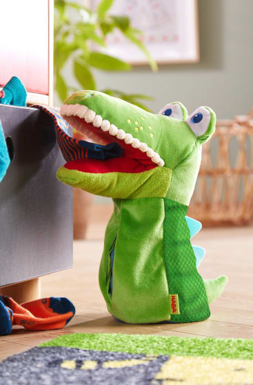 Eine grüne Plüschkrokodilpuppe mit blauen Stacheln hält eine gestreifte Socke im Maul und steht neben einer Schublade, in der weitere Socken zu sehen sind; im Hintergrund befinden sich ein Korb und eine Pflanze.